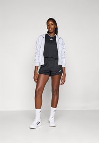 Tenue de sport noire avec un sweat à capuche zippé gris, ornée de trois rayures blanches, associée à des baskets blanches et des chaussettes, sur un fond neutre.