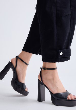 Pies con sandalias negras de plataforma y tacón alto con correas en el tobillo, combinadas con pantalones negros cortos con puños abotonados.