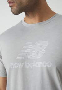 Siva majica kratkih rukava izrađena od glatkog materijala, s bijelim logotipom "New Balance" s paralelnim linijama i okruglim izrezom.