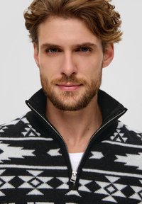 Schwarz-weißer gemusterter Reißverschluss-Pullover mit hohem Kragen, der geometrische Designs und strukturierten Strickstoff aufweist.