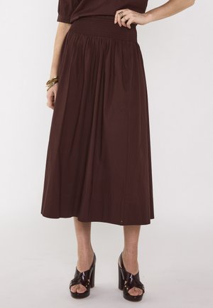 Mannequin portant une jupe midi taille haute marron foncé avec un tissu froncé et des sandales à talons hauts noires, accessoirisée de bagues et bracelets en or.