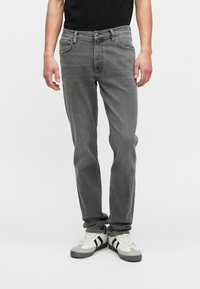 Homem a usar jeans cinzentos de corte reto e ténis brancos com riscas pretas, em pé contra um fundo claro e sem padrões.