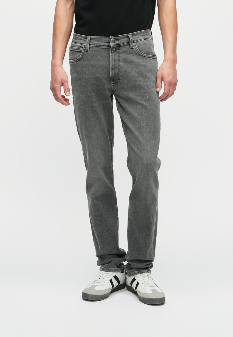 Homem a usar jeans cinzentos de corte reto e ténis brancos com riscas pretas, em pé contra um fundo claro e sem padrões.