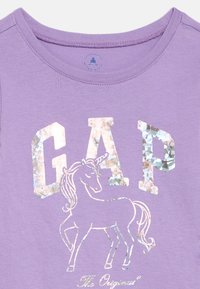 Levendula színű ing fényes ezüst "GAP" betűkkel, alatta ugráló unikornis illusztrációval, és az alján "The Original" szöveggel.