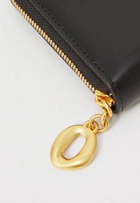 Portefeuille en faux cuir noir avec fermeture éclair et quincaillerie dorées, présentant un charme ovale doré distinctif attaché à la tirette de la fermeture éclair.