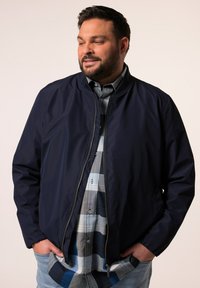 Marina bomberjacka med en slät yta och dragkedjestängning, lager över en ljusblå och grå rutskjorta. Avslappnad passform.
