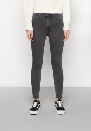 Persoon die nauwsluitende donkergrijze spijkerbroek, wit shirt met lange mouwen en zwart-witte sneakers draagt, staand tegen een effen witte muur.