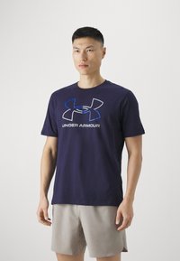 Tengerészkék, rövid ujjú pamut póló, amelyen elöl egy nagy szürke és kék Under Armour logó található, világos sorttal kiegészítve.