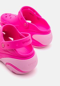 Fuchsia Crocs med en strukturerad design, utrustad med ventilationshål och en hälrem. Sulan är vit med rosa detaljer och varumärkeslogotyp.