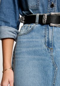 Zwarte leren riem met zilveren decoratieve gesp gedragen over lichtblauwe denim high-waisted jeans. Gerolde mouwen van een denim shirt zijn zichtbaar aan de bovenkant.