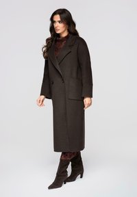 Abrigo largo de lana marrón con diseño de doble botonadura, grandes bolsillos delanteros y mangas anchas, usado sobre una camiseta burdeos con estampado y botas altas.