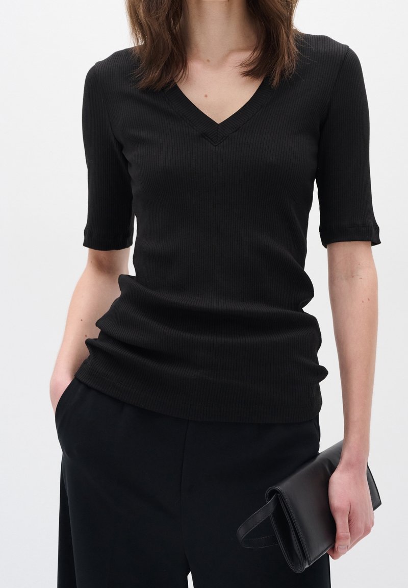 Svart ribbad stickad topp med V-ringning och veckade korta ärmar, kombinerad med svarta byxor och en svart kuvertväska. Enkelt mönster och textur.