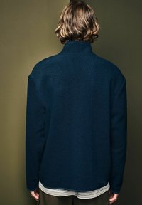 Marineblaue Fleecejacke mit hohem Kragen, überschnittenen Schultern und strukturierter Stoff. Kombiniert mit einem gestreiften Shirt, das am Saum hervorlugt.