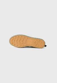 La semelle de la chaussure présente un design texturé avec une semelle extérieure en caoutchouc brun clair et une semelle intermédiaire gris foncé flexible ; elle comprend une petite languette noire.