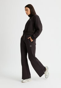 Sudadera negra corta con cuello alto, que presenta un logo bordado, combinada con pantalones anchos negros y zapatillas deportivas plateadas.