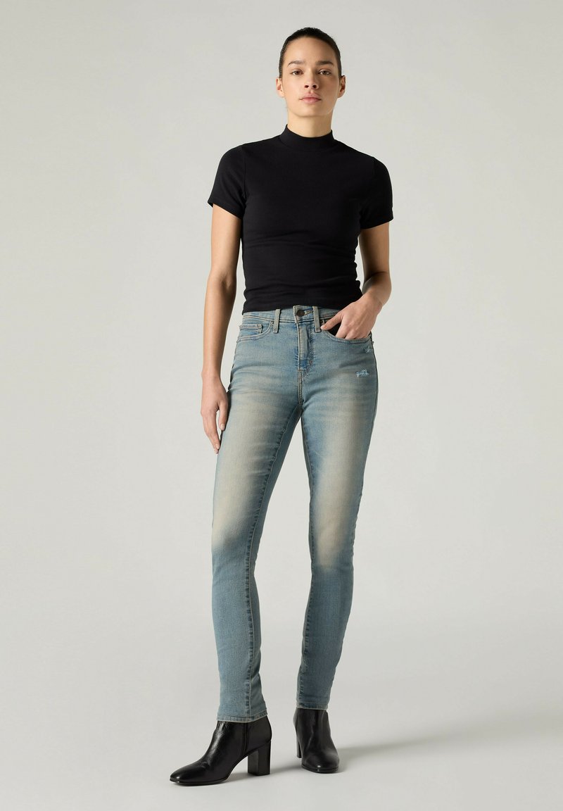Kvinde iført en sort kortærmet top, lys blå skinny jeans og sorte ankelstøvler med hæl står mod en ensfarvet lys baggrund.