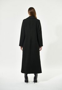 Lunga giacca nera con righe verticali, design sartoriale, ampio rever e spacchi laterali. Indossata sopra tacchi neri sottili, tessuto a trama liscia.