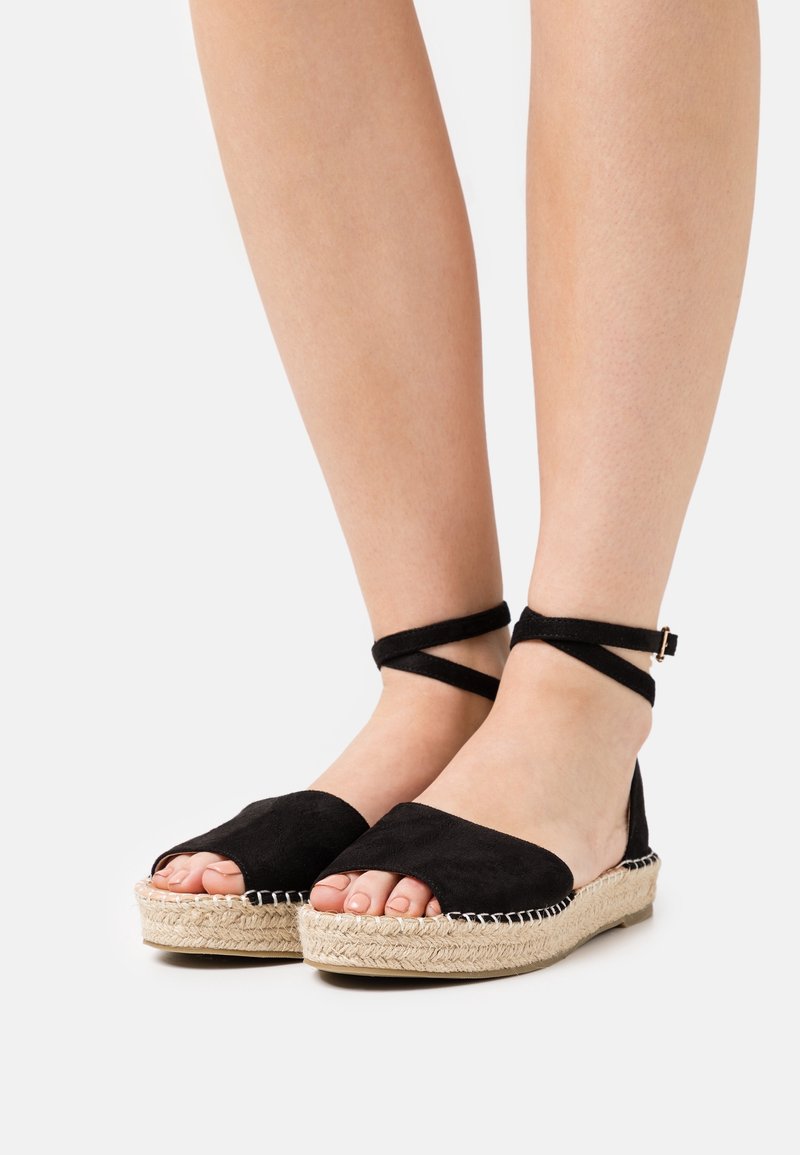 Pieds portant des sandales espadrilles noires avec des brides croisées autour de la cheville et des semelles compensées enveloppées de jute, présentées sur un fond clair uni.