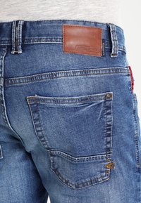 Jeans in denim di una tonalità blu media, con tasche posteriori, un'etichetta in pelle marrone sulla cintura e dettagli di cucitura a contrasto.