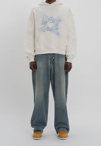 Sweat à capuche crème avec lettrage bleu, poche avant et capuche à cordon ; associé à un jean bleu délavé à coupe large et des chaussures marron.