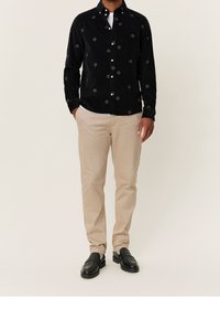 Camicia button-up in corduroy nera con motivi circolari ricamati, abbinata a pantaloni beige e scarpe nere, esposta su uno sfondo neutro.