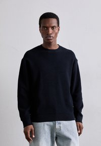 Marineblauer, gerippter Pullover mit rundem Halsausschnitt, lockerer Passform und langen Ärmeln, kombiniert mit hellblauen Jeans. Subtile Texturdetails sind sichtbar.