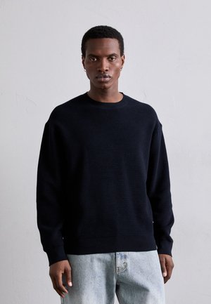 Pull à côtes bleu marine avec un col rond, coupe décontractée et manches longues, assorti à un jean en denim bleu clair. Détails de texture subtils visibles.
