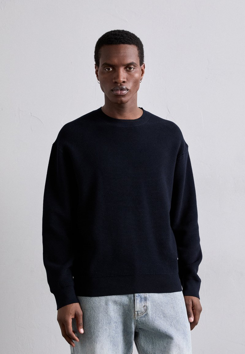 Marineblauer, gerippter Pullover mit rundem Halsausschnitt, lockerer Passform und langen Ärmeln, kombiniert mit hellblauen Jeans. Subtile Texturdetails sind sichtbar.