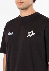 Must musta T-särk musta värviga, millel on valge tähe logo ja sinine tähe muster vasakul rinnal. Mugav lõige ümmarguse kaelaava ja lühikeste varrukatega.