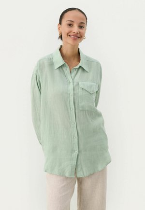 Cămașă verde deschis, cu nasturi, fabricată dintr-un material moale și ușor; are un buzunar frontal și o croială lejeră, cu guler.