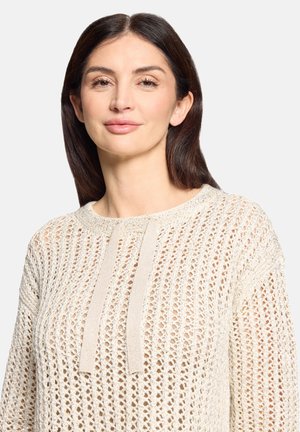 Femme aux cheveux raides et foncés portant un pull en crochet crème avec deux liens à l'avant, debout devant un fond clair uni.