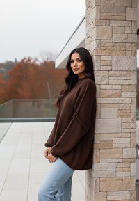 Brązowy sweter z fakturowanym splotem i oversize'owymi rękawami, zestawiony z jasnoniebieskimi dżinsami, oparty o kamienny mur na patio.