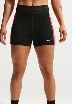 Primer plano de una persona que lleva puestos unos pantalones cortos deportivos negros de Nike con franjas laterales rojas y un logo blanco de Nike en la pierna izquierda.