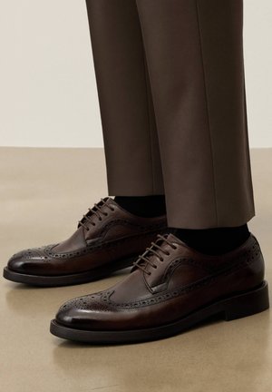 Tumši brūnas ādas brogues kurpes ar sīkiem perforējumiem, valkātas ar melnām zeķēm un brūnām kleitu biksēm uz pulēta grīdas seguma.