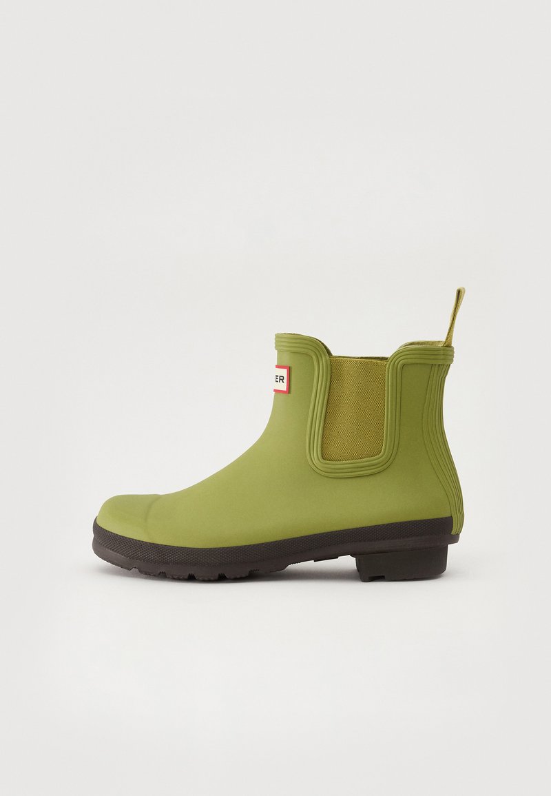 Bota de ankle verde em borracha com painéis laterais elásticos, sola texturizada castanha e uma alça na parte de trás. Superfície lisa com design de ponta arredondada.
