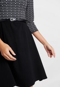 Robe noire avec une jupe ample et une taille cintrée, mettant en valeur un haut noir et blanc à motifs, accentuée par une fine ceinture noire.