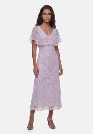 Frau trägt ein ärmelloses lavendelfarbenes Spitzenmidi-Kleid mit einer capeähnlichen Überlage und offene Lavendel-Absatzschuhe mit dünnen Riemchen.