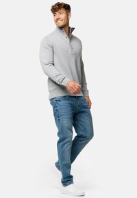 Jersey gris con cremallera de cuarto, puños acanalados y dos botones, combinado con jeans azules y zapatillas blancas, con fondo blanco.