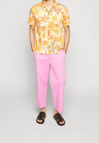 Camicia gialla a maniche corte con stampa floreale e bottoni, abbinata a pantaloni rosa a gamba larga e sandali neri con cinturini.