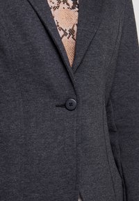 Blazer gris oscuro con solapa con muesca, cierre de un solo botón y un patrón tejido texturizado debajo. Presenta un sutil estiramiento y un ajuste entallado.