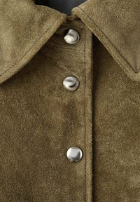 Chemise en daim vert olive avec col, ornée de boutons métalliques argentés sur le devant. Texture douce et détails de couture subtils.