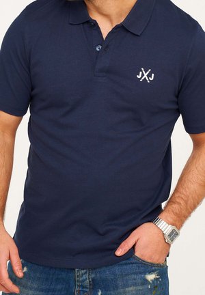 Mand iført navyblå kortærmet polo med et krydset stokke-logo på brystet, slidte blå jeans og sølvfarvet ur på venstre hånd.