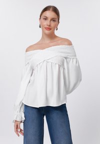 Weiße Bluse mit schulterfreiem Schnitt, mit Raffung, langen Ärmeln und gerafften Abschlüssen. Fließende Silhouette aus einem weichen, leichten Stoff.