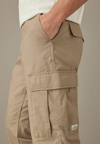 Beige cargo bukser med tekstureret stof, sidelommer og spændebånd. Har flere cargo lommer og et vævet mærke.