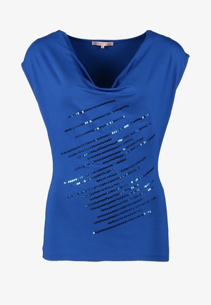 Kvinde iført en blå ærmeløs top med skinnende diagonale striber, sorte smalle bukser, sorte ankelstøvler med hæl, der holder en sort clutch.