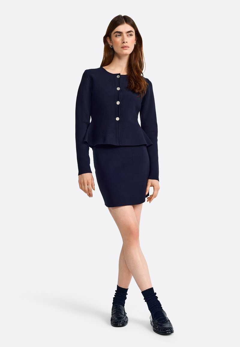 Conjunto de dos piezas en navy que incluye un vestido ajustado y una chaqueta peplum con botones plateados, complementado con mocasines negros y calcetines hasta los tobillos.