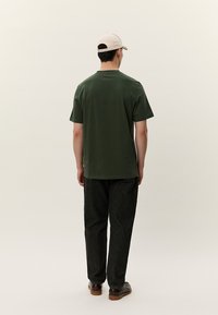 Zelená košile s krátkým rukávem, volný střih, v kombinaci s tmavými texturovanými kalhotami a hnědými botami. Model má na sobě světlou kšiltovku.