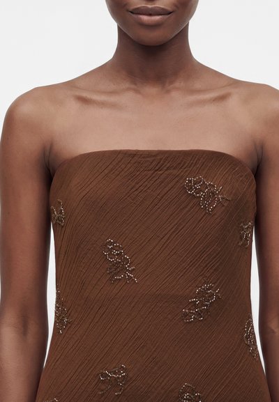 Femme portant une robe marron sans bretelles en tissu texturé avec de petits groupes de perles décoratives en forme de nœuds à l'avant.
