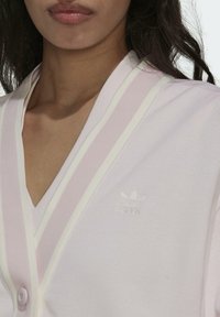 Ljusrosa cardigan med en texturerad tyg, prydd med vita ränder längs kanterna och en liten, vit adidas-logotyp på bröstet.
