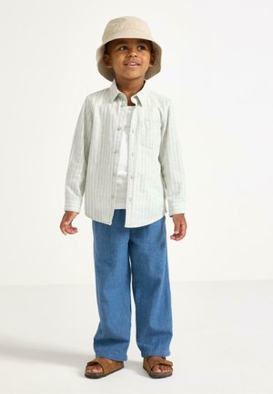 Ung dreng smilende, iført beige bøllehat, stribet skjorte med knapper, hvid T-shirt, blå bukser og brune sandaler, stående på hvid baggrund.